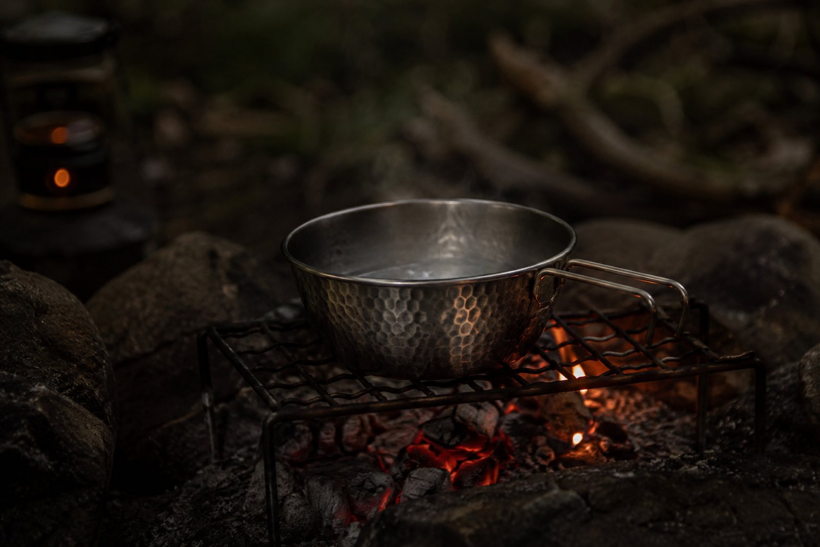 A titanium sierra cup on a fire grate above glowing coals with thin steam rising
